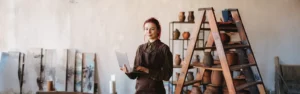 A female painter with red hair, wearing a dark green shirt and a brown apron, holds a laptop and poses in her art studio with sculpted pitches and jars on a shelf and various canvases in the background.