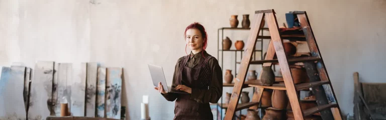 A female painter with red hair, wearing a dark green shirt and a brown apron, holds a laptop and poses in her art studio with sculpted pitches and jars on a shelf and various canvases in the background.