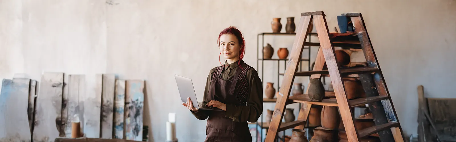 A female painter with red hair, wearing a dark green shirt and a brown apron, holds a laptop and poses in her art studio with sculpted pitches and jars on a shelf and various canvases in the background.