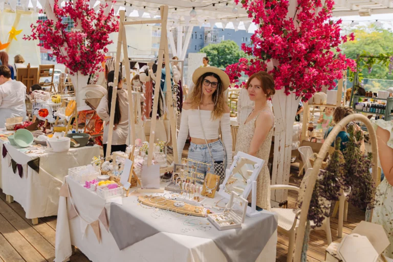 Craft vendors smile and stand behind their jewelry craft table at a craft market decorated with neutral-tone decor, pink bougainvillea, and boho crafts.