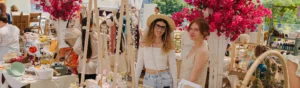 Craft vendors smile and stand behind their jewelry craft table at a craft market decorated with neutral-tone decor, pink bougainvillea, and boho crafts.
