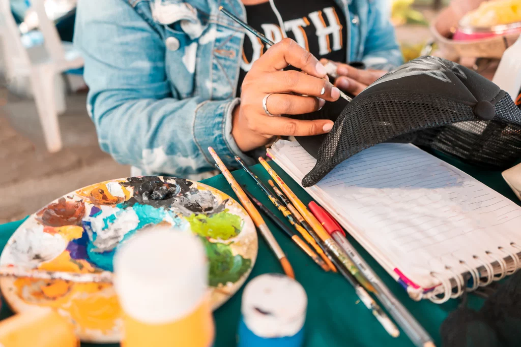 A woman wearing a black graphic tee and a denim shirt paints on a black trucker hat at a table covered with green cloth. On the table are a white notebook, paintbrushes, a palette, and paint bottles.