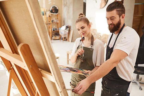 A young bearded man teaches a young woman more about how to paint as they smile and look at her canvas in a sunny studio.