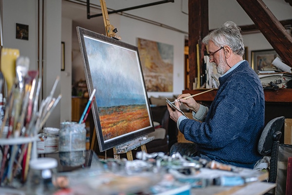 An older man sits in his home studio and cleans his brush as he finishes a painting of a peaceful field landscape.