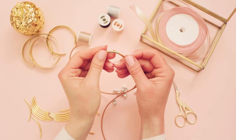 A pair of hands are crafting handmade gold jewelry on a pink background.