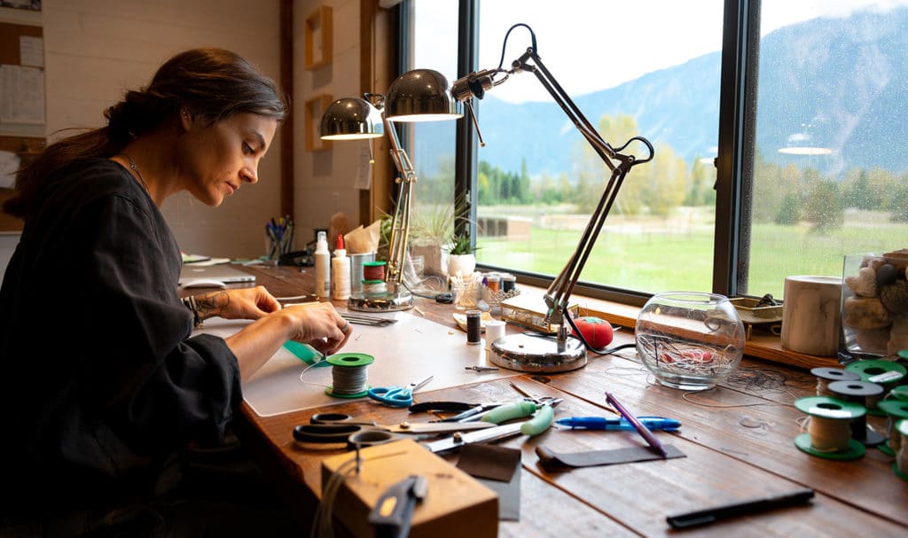 A woman works on some jewelry at her desk next to a window that looks out at mountains and a grassy field. She is using the light of a lamp to get a better look at the smaller details in her work.