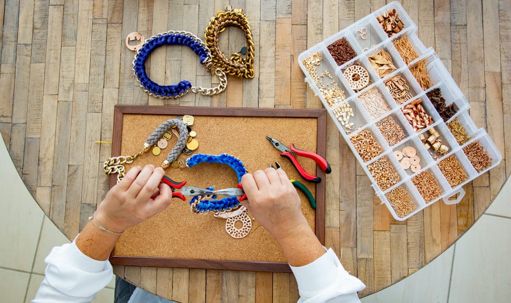 An above view of a pair of hands, a mat with partially completed jewelry, jewelry tools, a box of different beads, and two finished necklaces all rest on a round wood table.