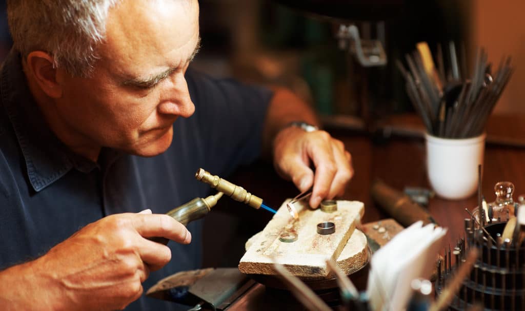 An older man uses a small jewelers torch and pliers to work on constructing some rings inside his jewelry studio.