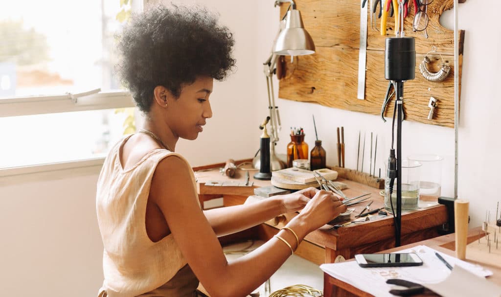 A young woman sits in her home jewelry studio as she handcrafts her next piece on a desk full of jewelers tools and supplies.