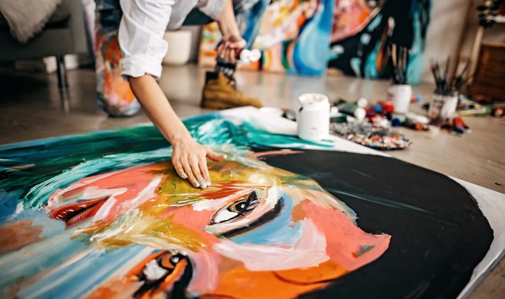 An artist kneels on the concrete floor over a giant portrait of a woman as they hand paint in their studio.