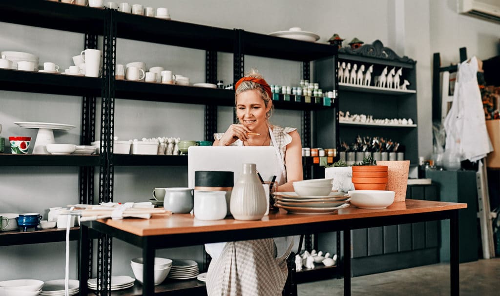 A woman sits in her home art studio at a table looking at her computer for etsy product liability insurance. Her pottery she sells online sits on shelving behind her insured by ACT.