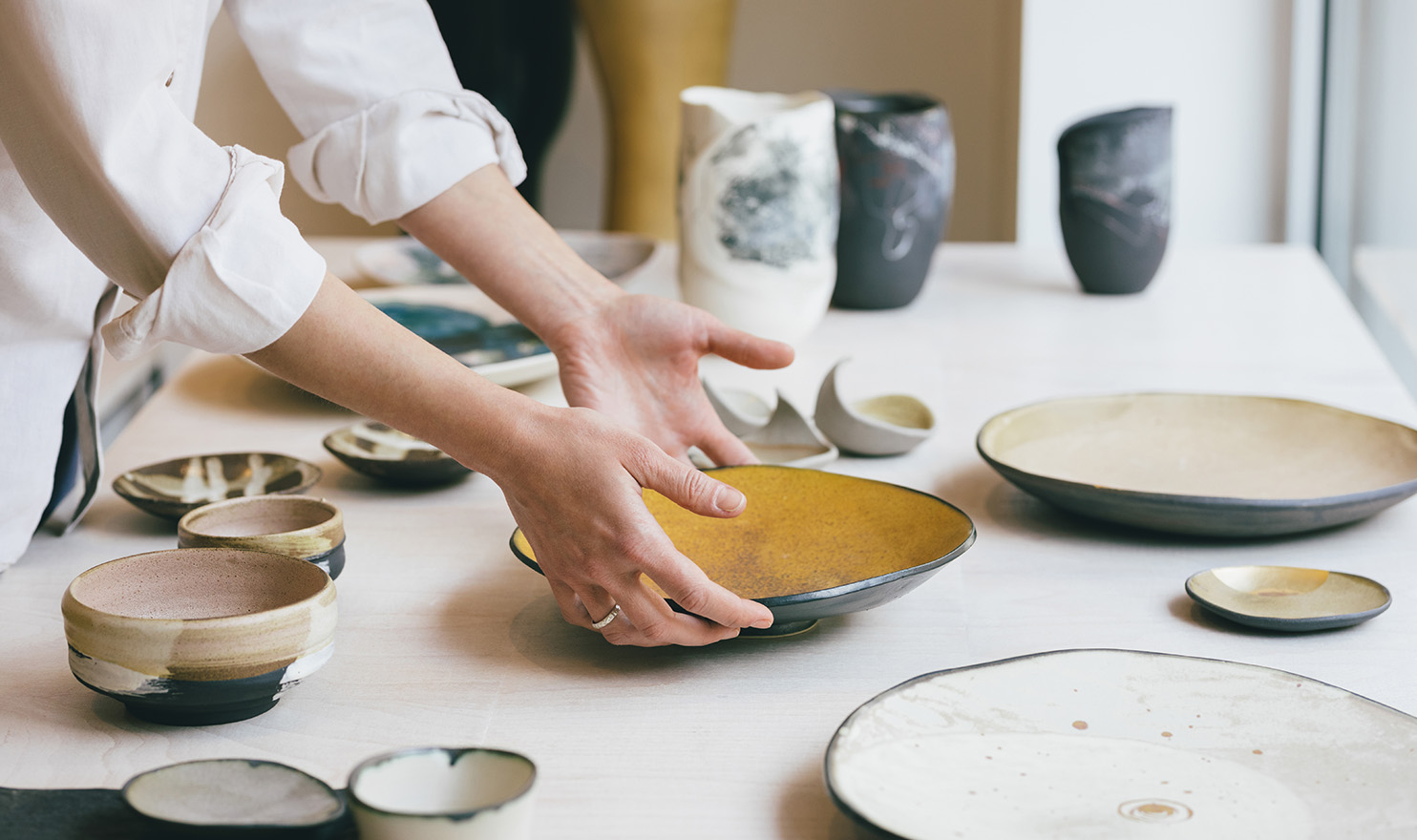 A woman's hands are carefully setting down a handmade plate on a white table next to other pieces of handmade pottery.