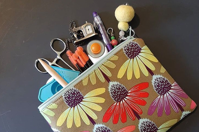 A brown handbag with a colorful daisy pattern lies on a gray background and has several unique sewing tools falling out of it.