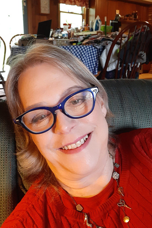 A photo of Joan Radell in a red blouse and glasses in her home.