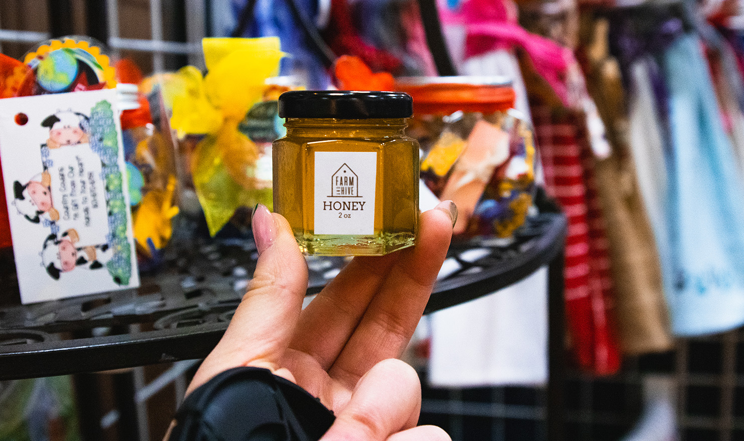 An image of a small pot of honey being held up with a booth at Holy Cow Boutique in the background.