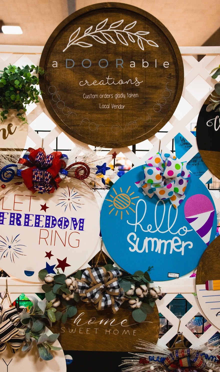 Round handmade wooden door signs are hung on a white trellis in the aDOORable creations booth.