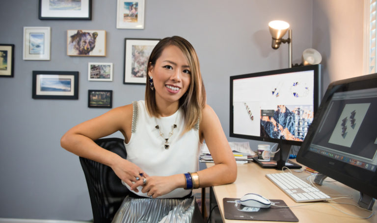 Hsiang-Ting Yen poses and smiles at her desk where she makes graphic mock-ups of jewelry on her computer.