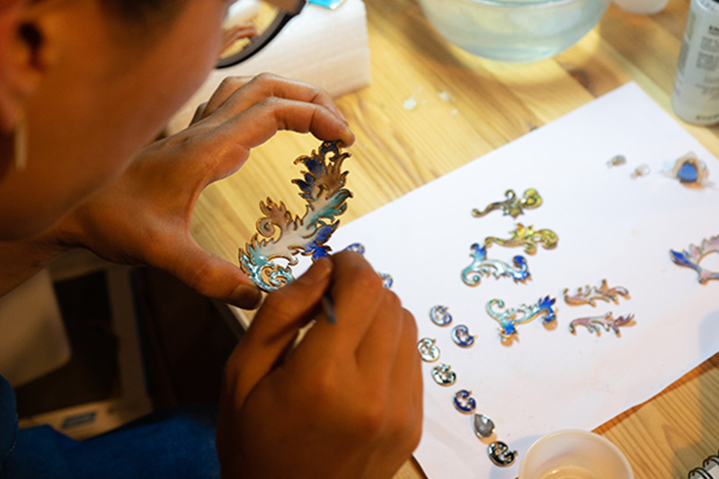 Hsiang-Ting Yen working on a set of custom jewelry. She is holding a unique pendant that has swirls of blue and white color as she adds additional charms and jewels to it.
