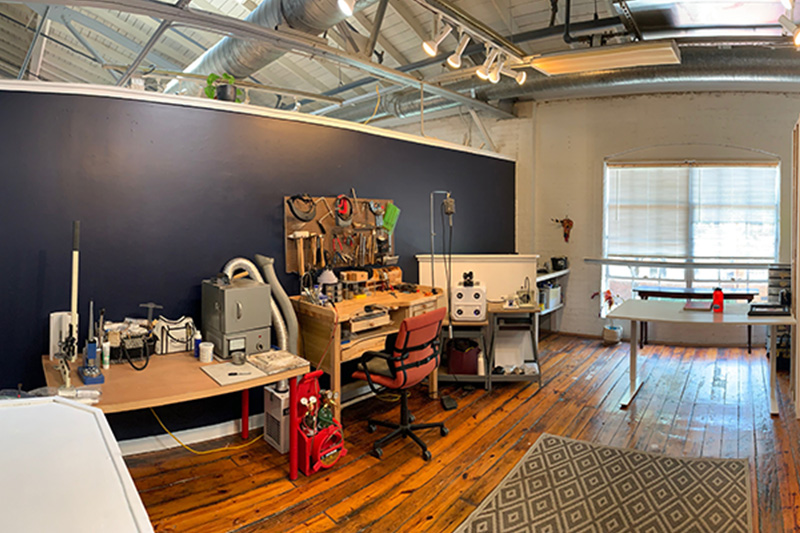A panoramic shot of a jewelers workspace, complete with tools, a small kiln, a drawing board, and a lot of tabletop space to create custom pieces.