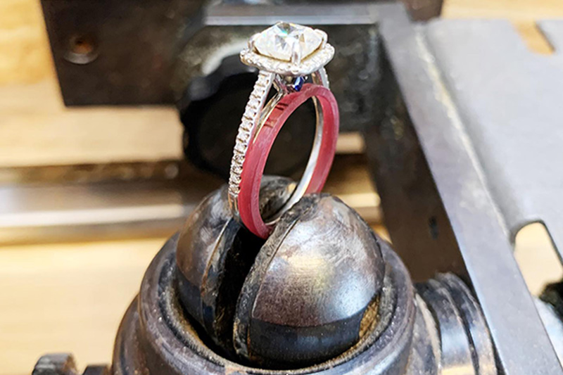 A handcrafted silver ring with a square cut diamond on a cushion of small diamonds sits in a special jewelers tool to prop the ring upright against a mold for the wedding band.