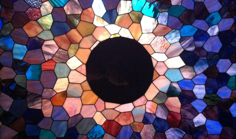A stained colorful stained glass depiction of a solar eclipse with a light shining through the glass.