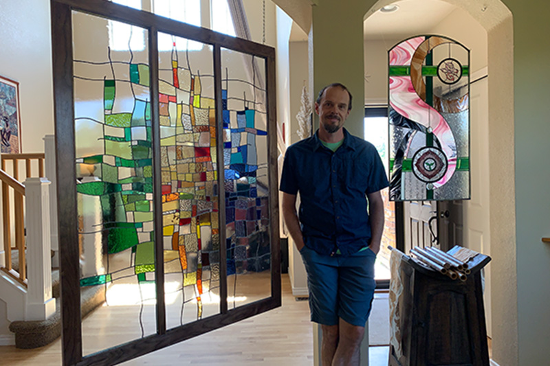 Jeremy Walker in a short sleeve blue shirt and denim shorts poses with his hands in his pockets while standing between two large stained glass installations he made and has hanging in his home.