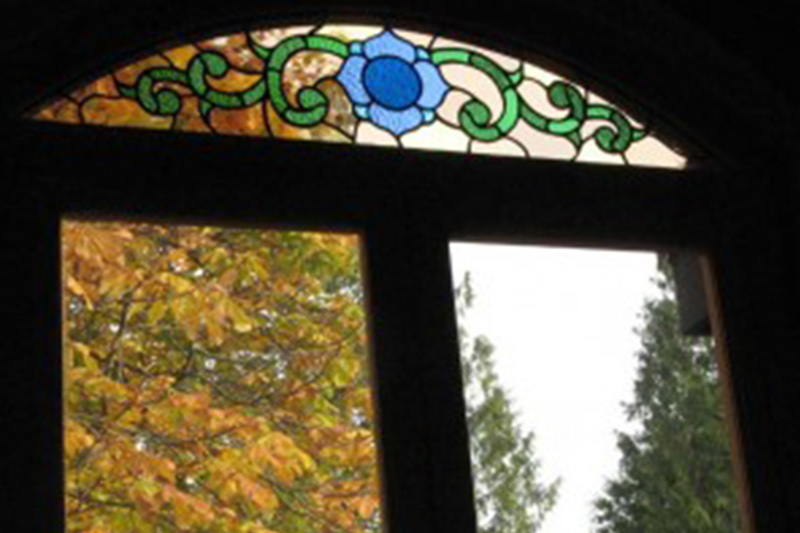 A window arch above a set of two windows has a small stained glass design of a blue flower in the center and green vines stemming out from it.