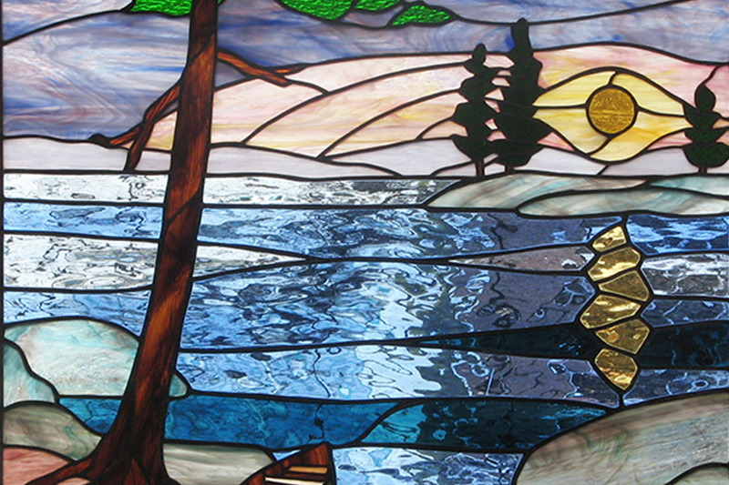 A nature scene of a lake, a setting sun, and pine trees are depicted in a stained glass window.