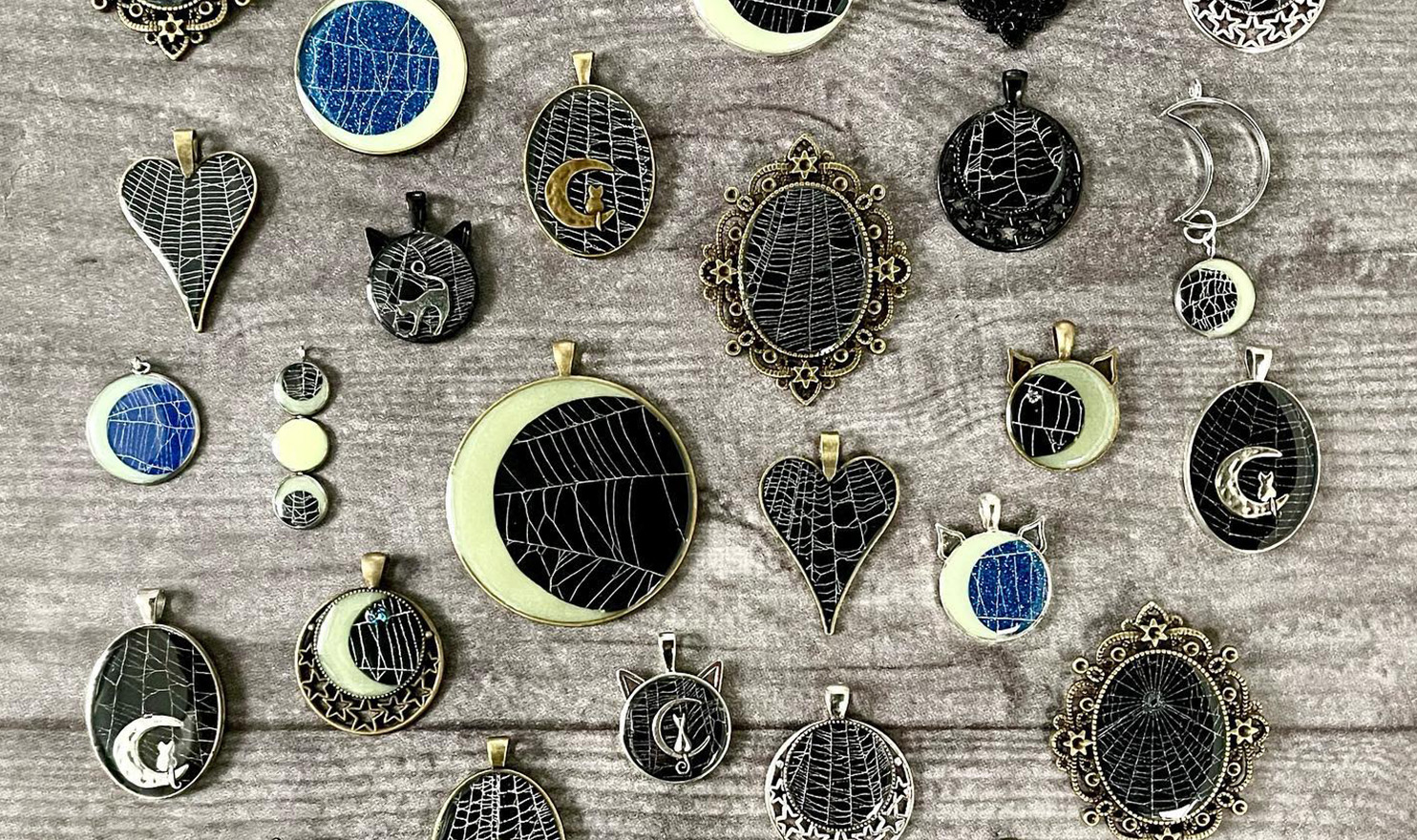 A variety of gothic inspired necklace pendants laying on a wood table feature preserved spiderwebs on them.