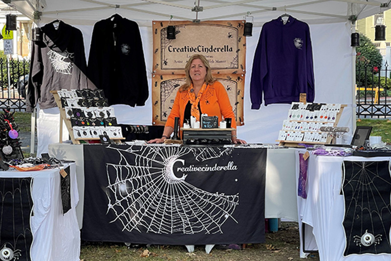 Laura Morriseau standing at a table in her event booth surrounded by her handcrafted items, such as shirts and jewelry.