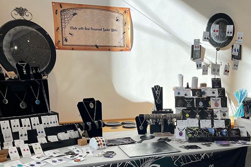 A variety of handcrafted spiderweb products sit neatly organized on a table to be sold at an event.