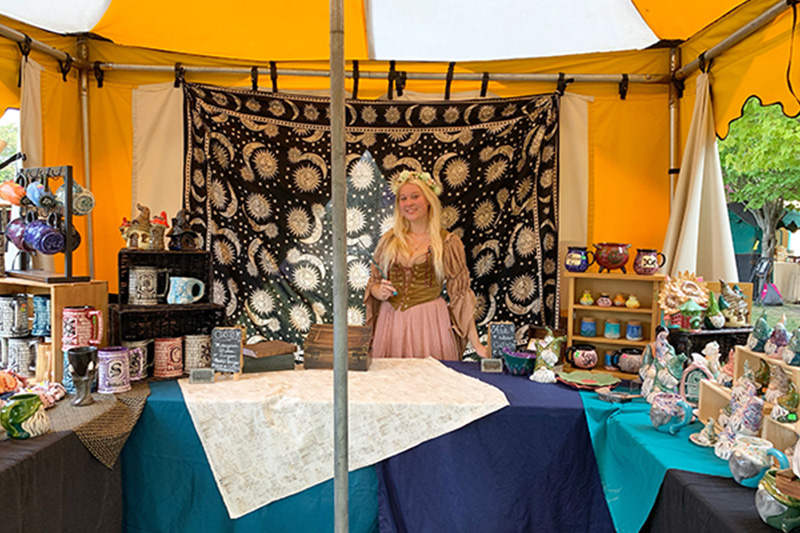 Marika dresses in to the renaissance theme she is attending poses in her booth full of hand painted pottery she plans to sell at the event.