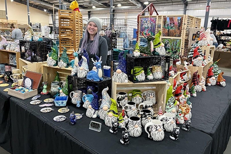 Marika poses behind a table full of displays with her hand painted pottery at a local event.