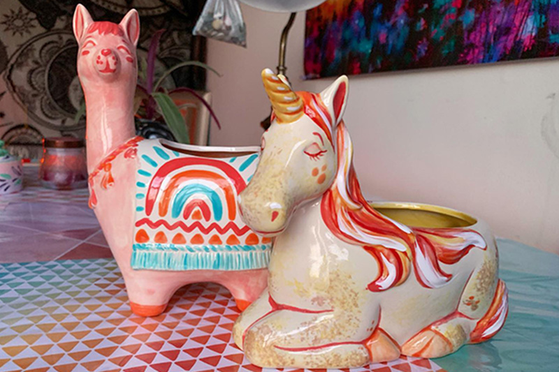 Two pieces of hand painted pottery, a llama and a unicorn, are posed next to one another on a table for Marika to photograph for her social media channels.