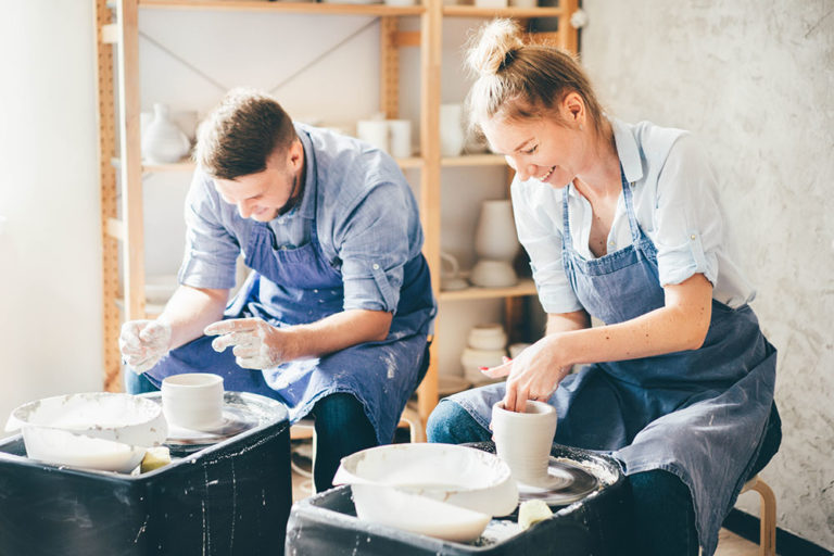 Two potters are happily working on their pieces side by side in a pottery studio.