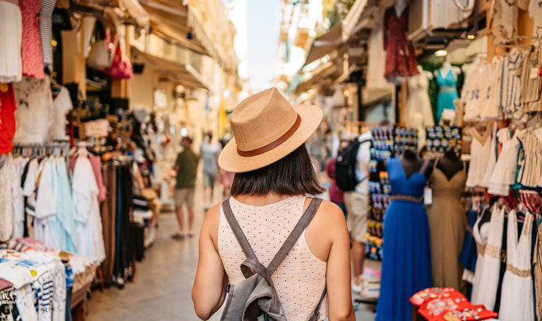 A woman's back is to the camera as she walks through the aisle of an outdoor market where vendors are selling items from their booths and tents.