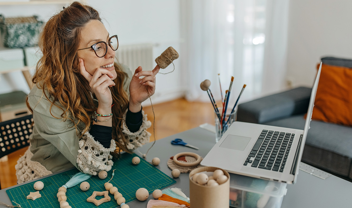 A woman sits in her home craft studio working on a laptop next to a craft project she is working on.