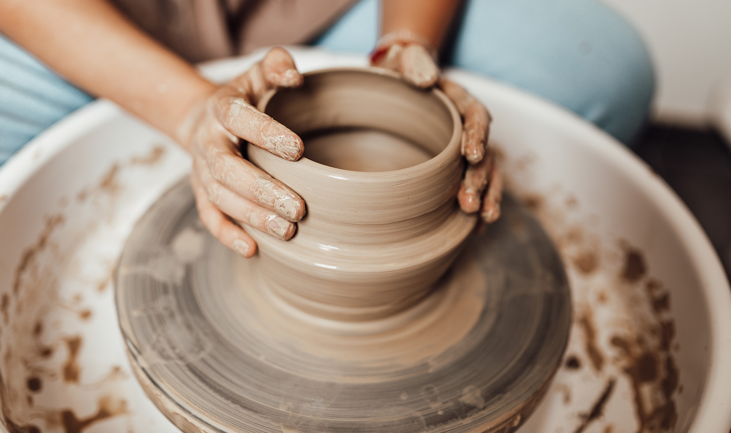 A potter's hands are shaping and molding clay on a wheel into a pot.