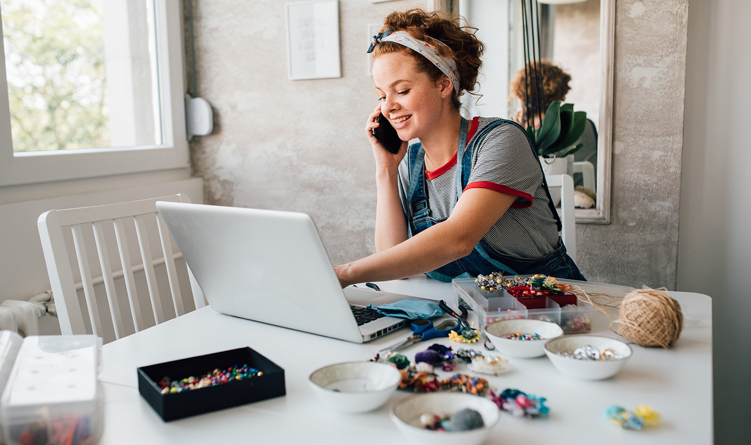 A young craft jeweler is speaking on the phone to her agent about product liability insurance as she works on her pieces next her laptop on a white table in her beige colored home office.