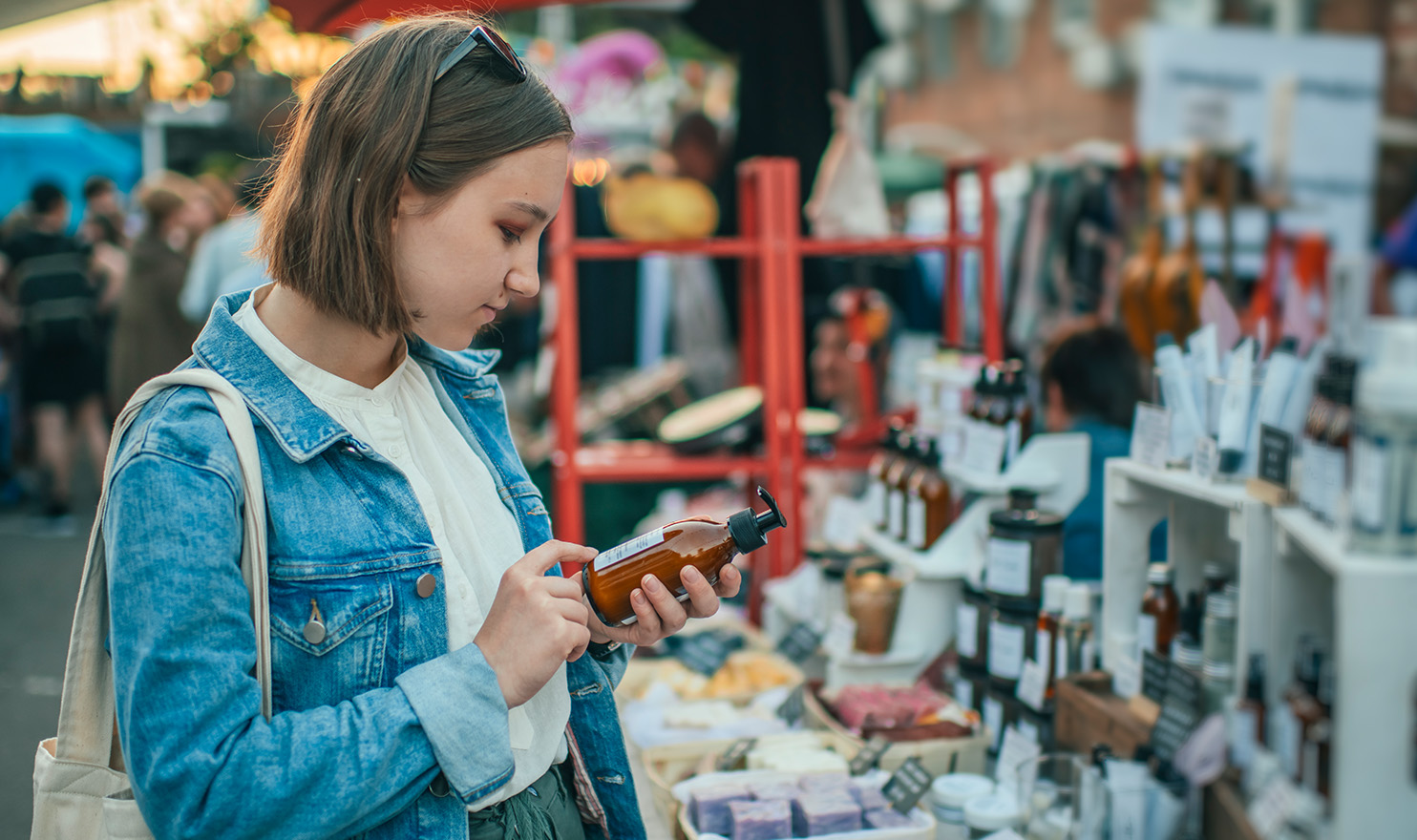 A young woman is looking at the ingredients and reading the label on a bottle of handmade lotion at a craft fair.