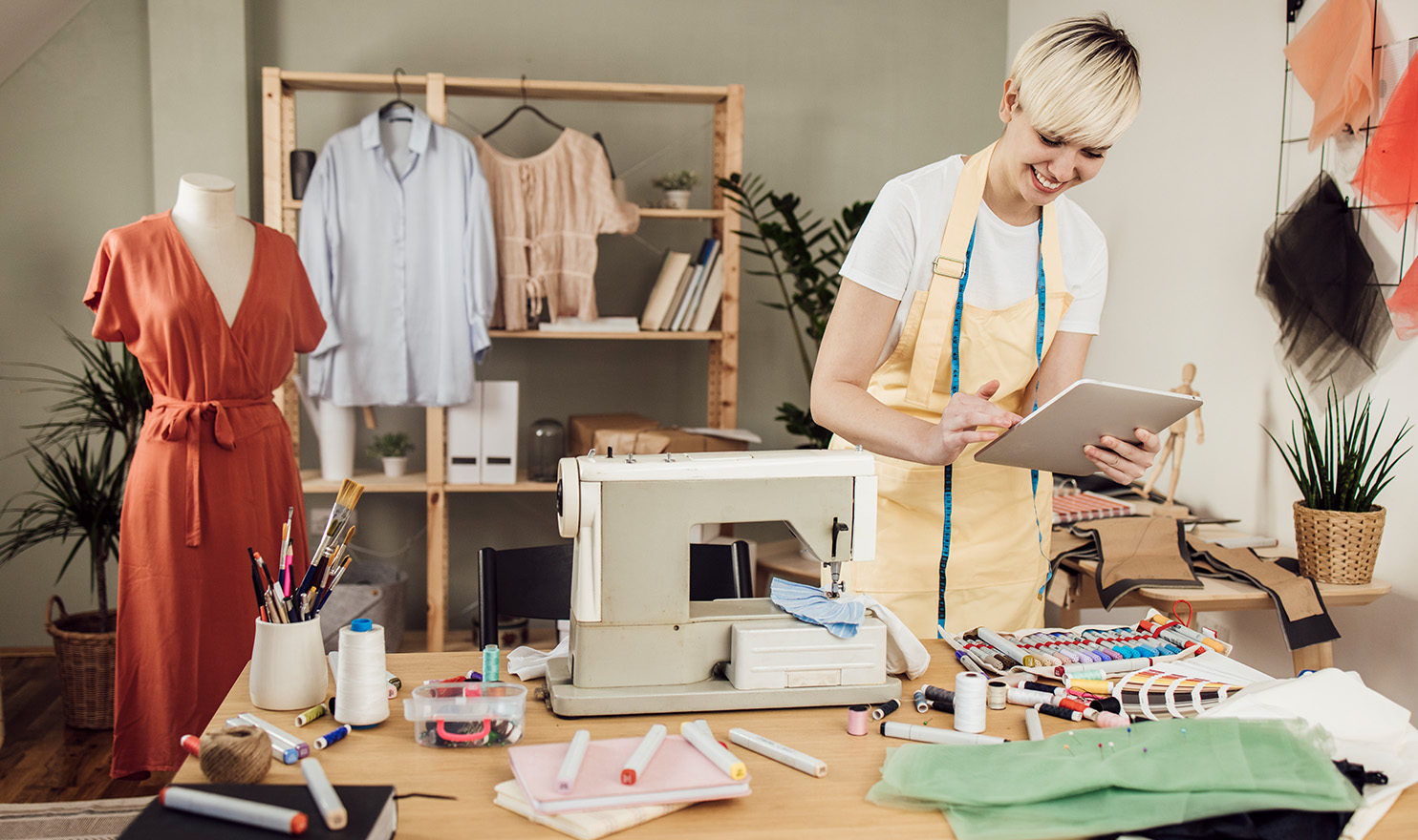 A seamstress is working in her home studio and is purchasing product liability insurance on her electronic tablet while standing next to her sewing table full of supplies.