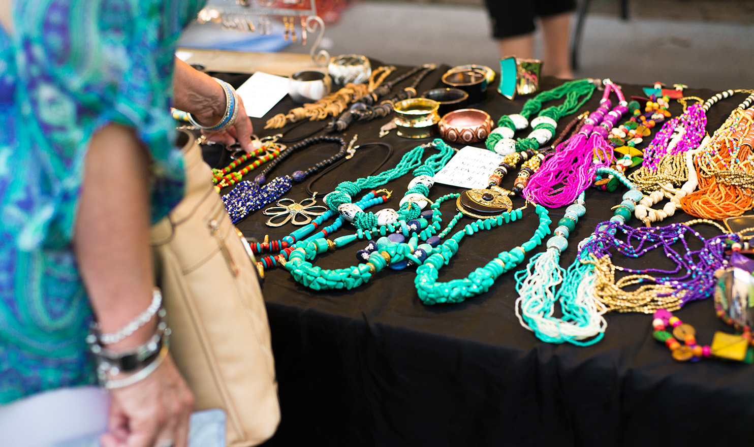 An event goer is looking at handmade necklaces out on display on a black table in a vendor's booth.