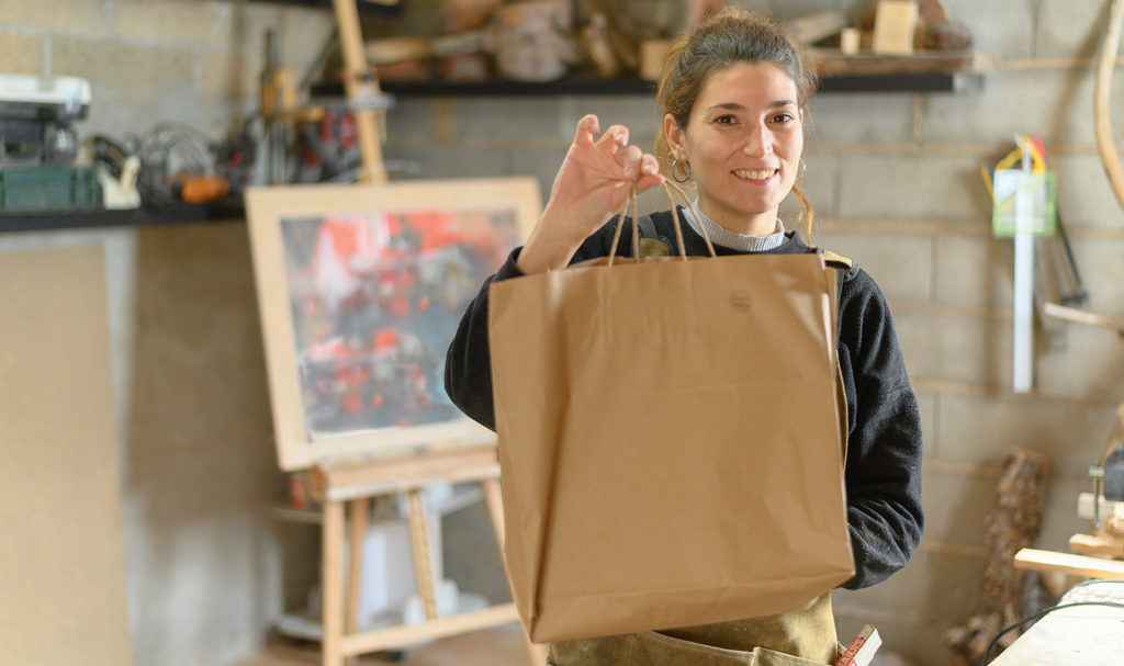 A vendor is smiling with a brown paper bag that she has filled with a customer's purchases.