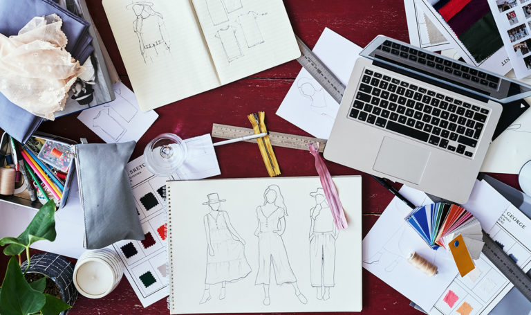 An above view of a laptop on a desk amongst different items and sketches for fashion design.