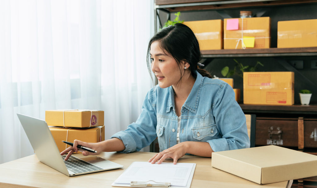 A young business owner is in her home office organizing emails on her laptop next to packages ready to send to her customers.