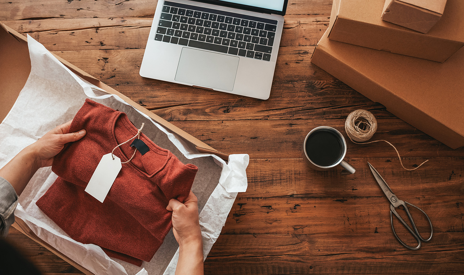 A small creative business owner is packing up a handmade shirt into a box atop their workstation next to a laptop and supplies.