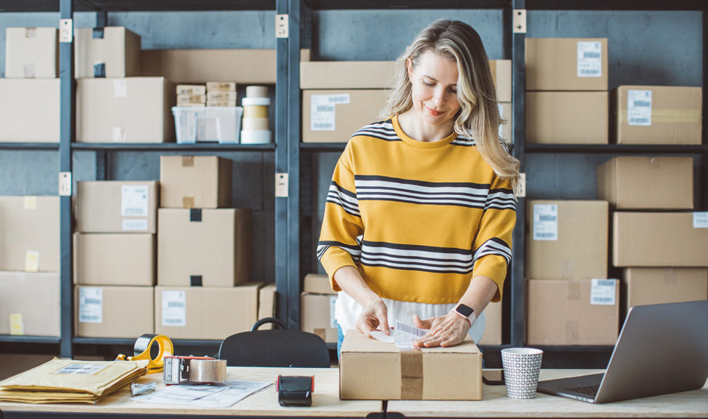 A small business owner is placing a shipping label on a package in her workspace.