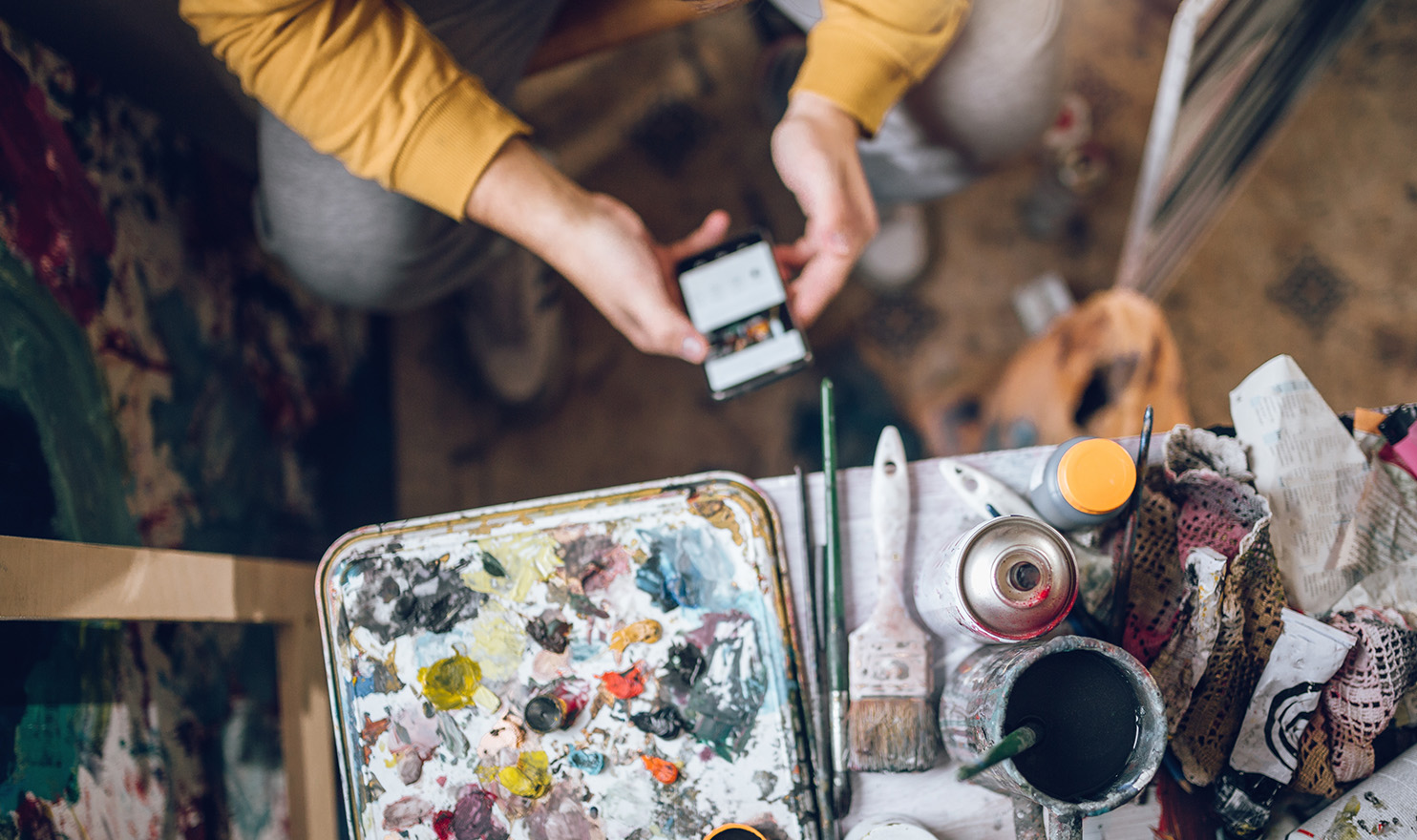 An overhead shot of an artist on their phone in their studio next to their painting supplies.