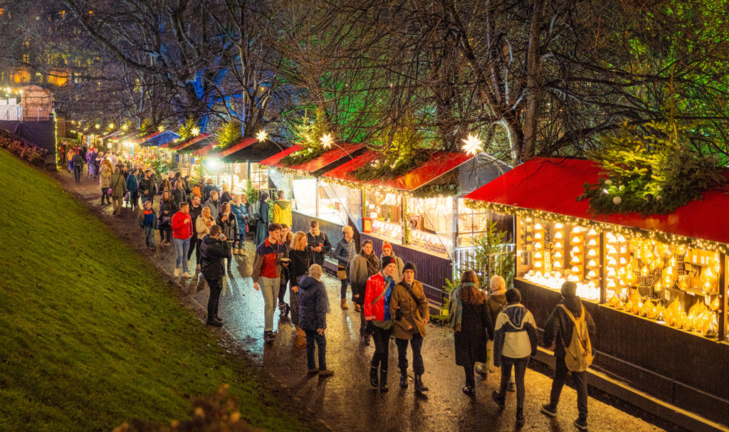 An outdoor evening holiday market is in full swing as the booths are lit up with Christmas lights and customers are walking from booth to booth.