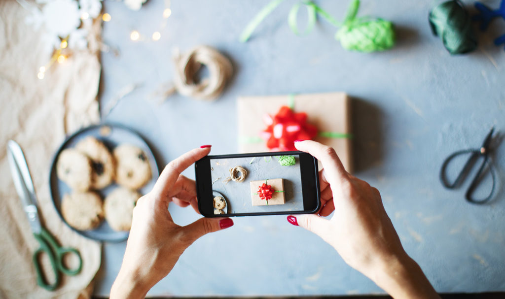 An overhead shot of a person taking a photo on a smart phone of their new holiday wrapping for a gift. This will be a special holiday post for their business social media pages.